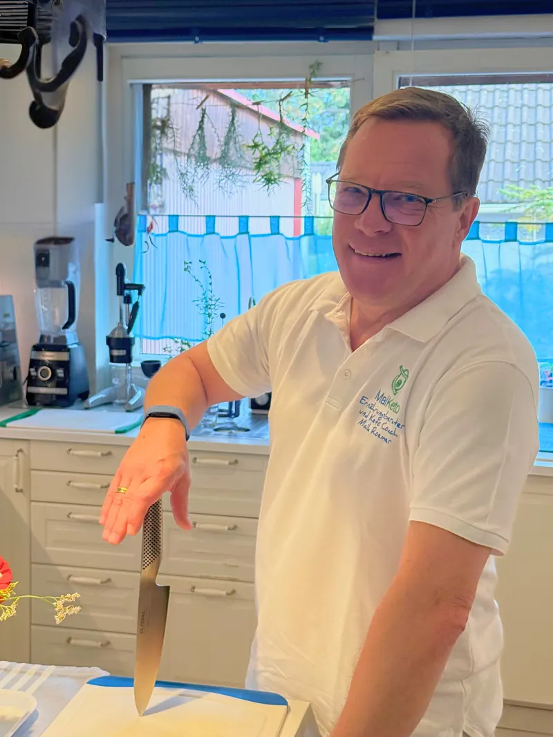 Maik Roemer lacht in seiner Küche, die Hand über einem Kochmesser auf dem Schneidebrett